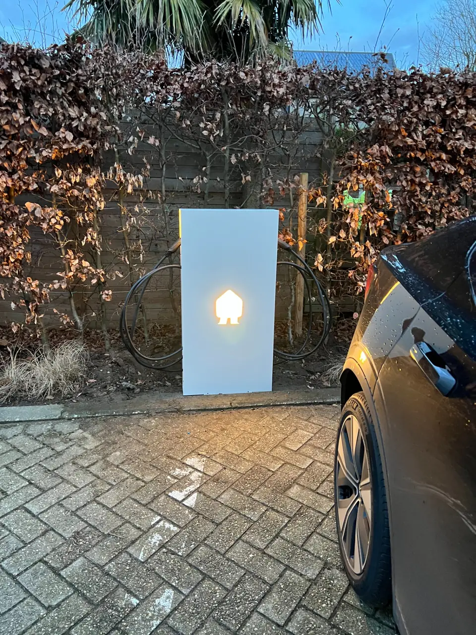 Verlichte laadpaal op een oprit bij avond, naast een elektrische wagen